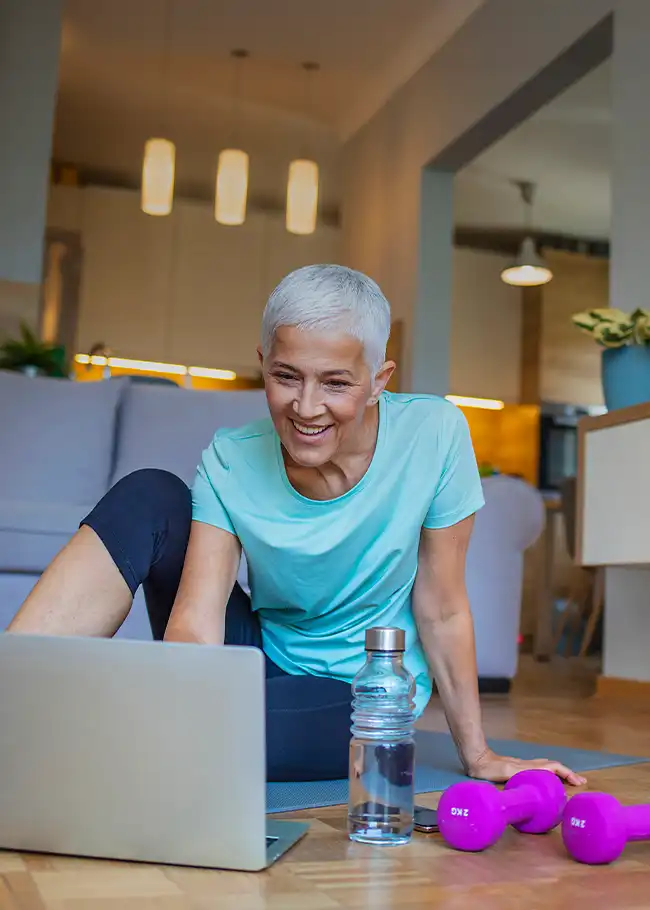 Gray-haired woman in teal top smiling at a laptop on living room floor with water bottle and pink dumbbell 