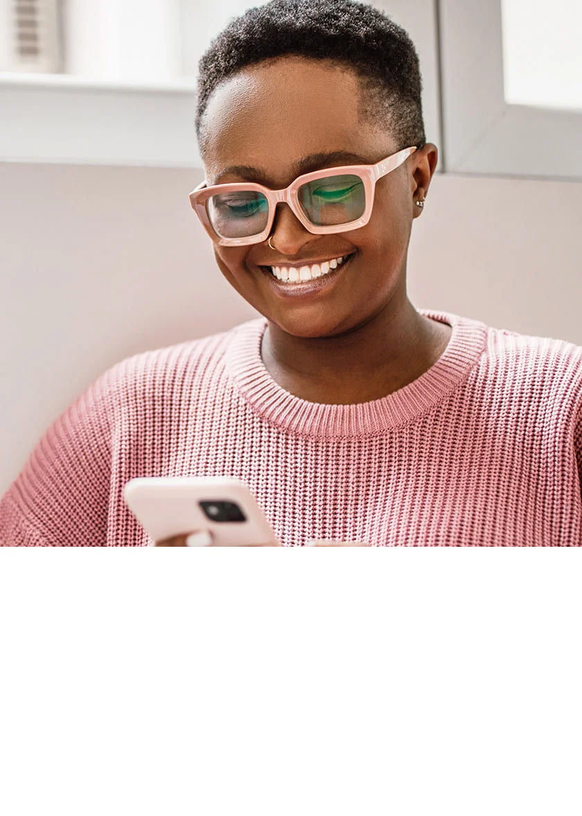 Woman in glasses and coral sweater smiling while looking at her smartphone