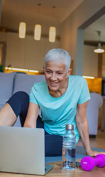 Gray-haired woman in teal top smiling at a laptop on living room floor with water bottle and pink dumbbell