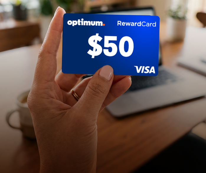 A hand holds a blue Optimum $50 Visa RewardCard over a blurred desk with a plant behind