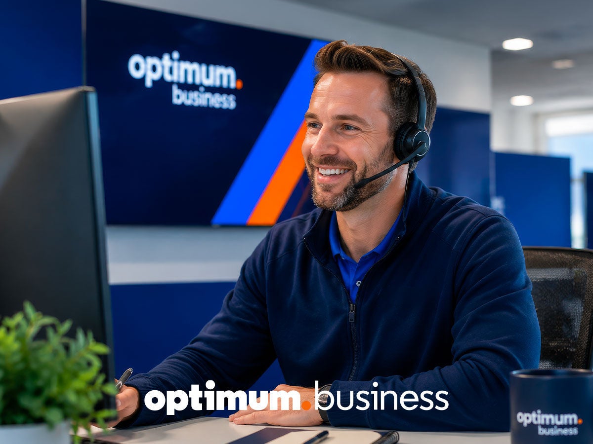 Smiling Optimum Business customer support representative wearing a headset at a desk, with the Optimum Business logo on screen behind him.