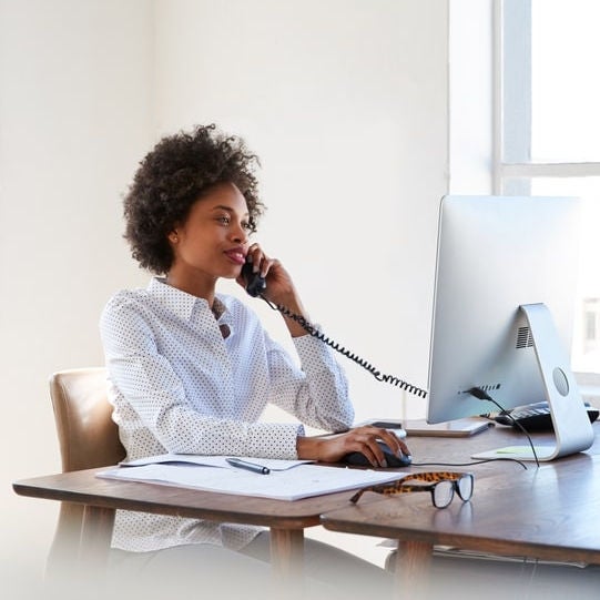 Una mujer profesional en un entorno de oficina, hablando por teléfono en su escritorio, junto a una computadora de escritorio. La escena destaca un entorno de trabajo moderno con amplios ventanales y luz natural, lo que sugiere una comunicación eficiente a través de un sistema troncal PRI.
