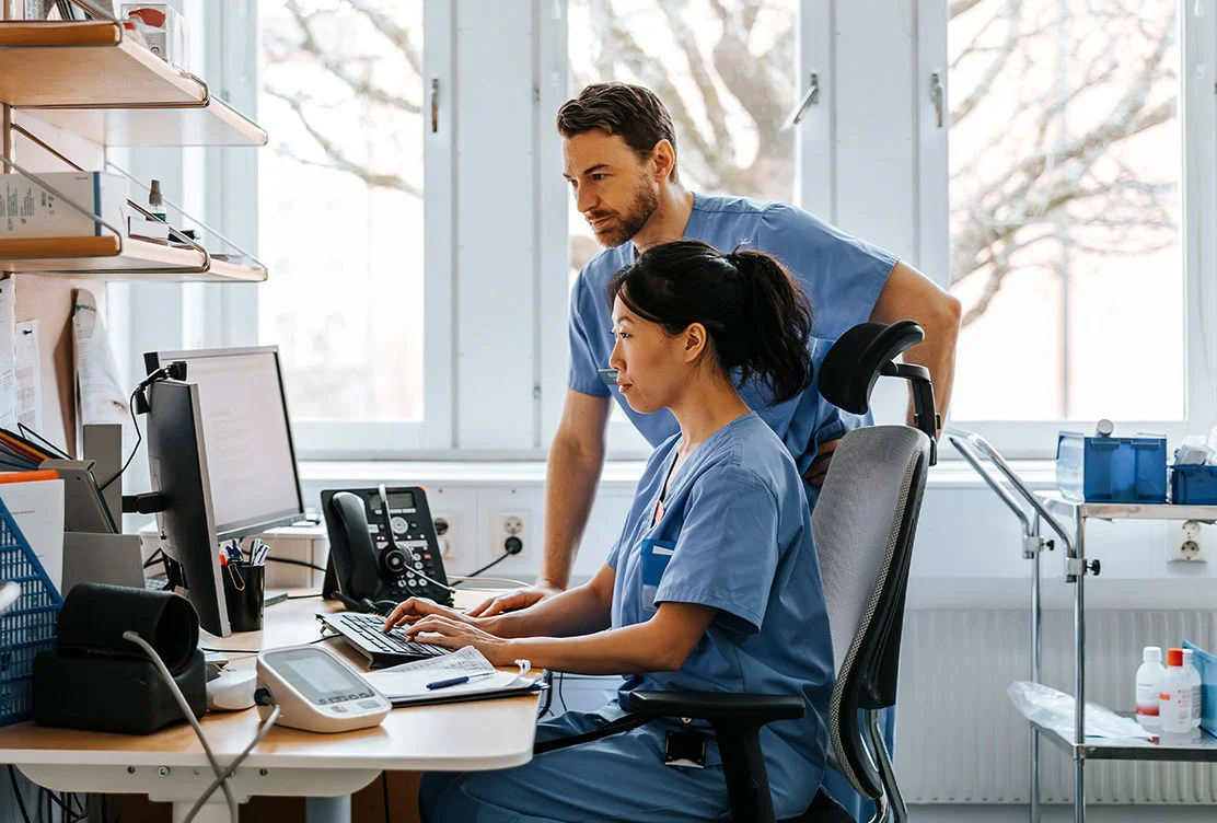 Healthcare worker using a desktop computer at a nurses station with a colleague standing behind 
