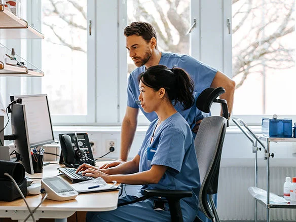 Healthcare worker using a desktop computer at a nurses station with a colleague standing behind 