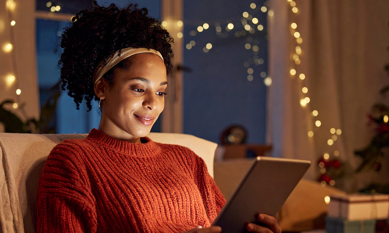 Smiling woman in orange sweater holding tablet, warm string lights in dark room behind her