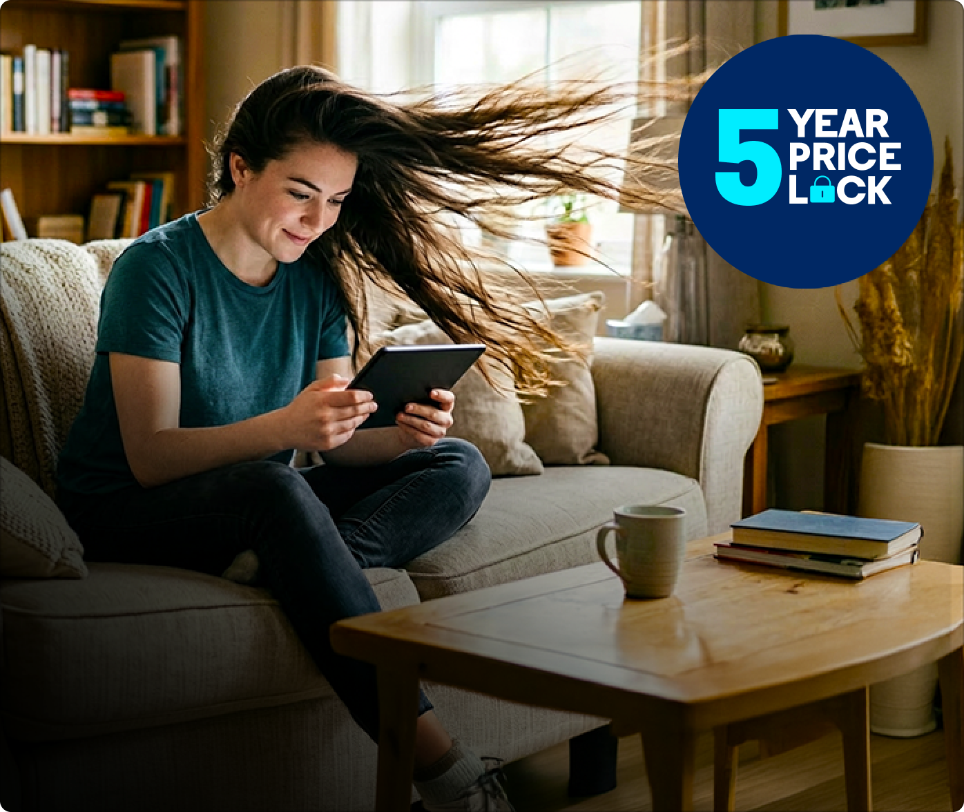 Woman with long flowing hair sits on a sofa looking at a tablet, smiling; coffee mug and books on the table