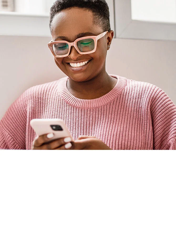 Woman in glasses and coral sweater smiling while looking at her smartphone
