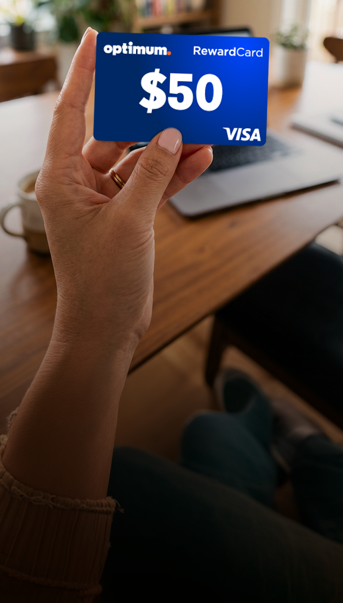 A hand holds a blue Optimum $50 Visa RewardCard over a blurred desk with a plant behind