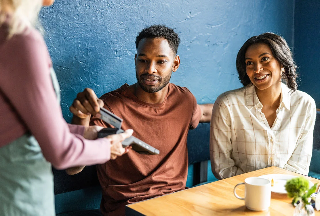 Two people exchanging a credit card payment across a restaurant table while smiling 