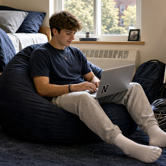 College student in a dorm room typing on a laptop while seated on a navy bean bag chair