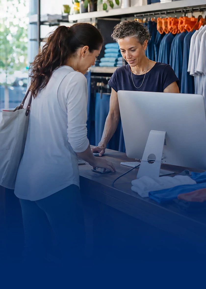 Two women looking at a desktop computer screen inside a clothing boutique with garment racks in the background