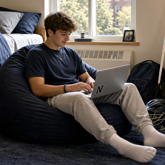 College student in a dorm room typing on a laptop while seated on a navy bean bag chair
