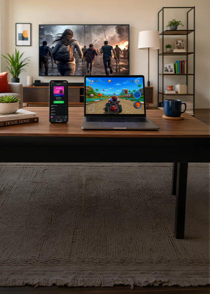 Mesa de centro en la sala de estar con un teléfono inteligente y una computadora portátil que muestran un juego de carreras, frente a un televisor que reproduce una escena de acción