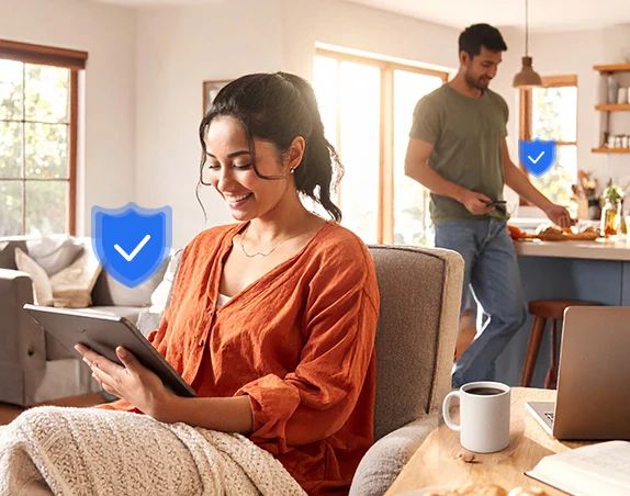 Mujer con cárdigan color óxido sentada en un sofá usando un teléfono inteligente, hombre relajándose detrás de ella, comida sobre la mesa, insignia de verificación azul