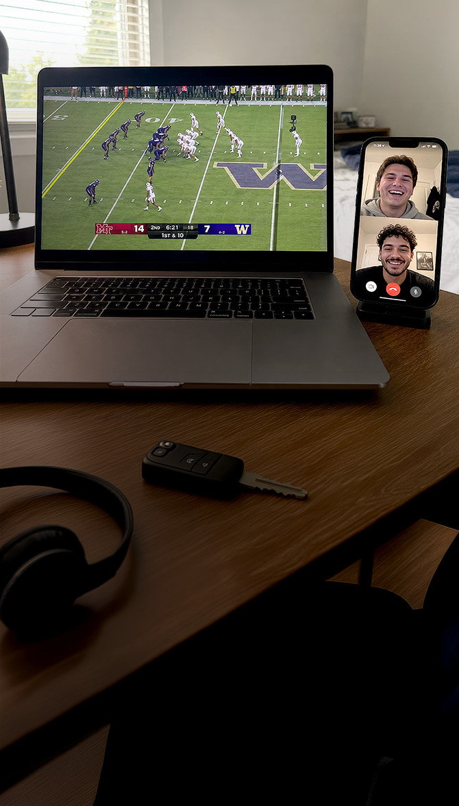Computadora portátil que muestra un partido de fútbol americano junto a un teléfono inteligente en videollamada con dos hombres sonrientes, sobre un escritorio