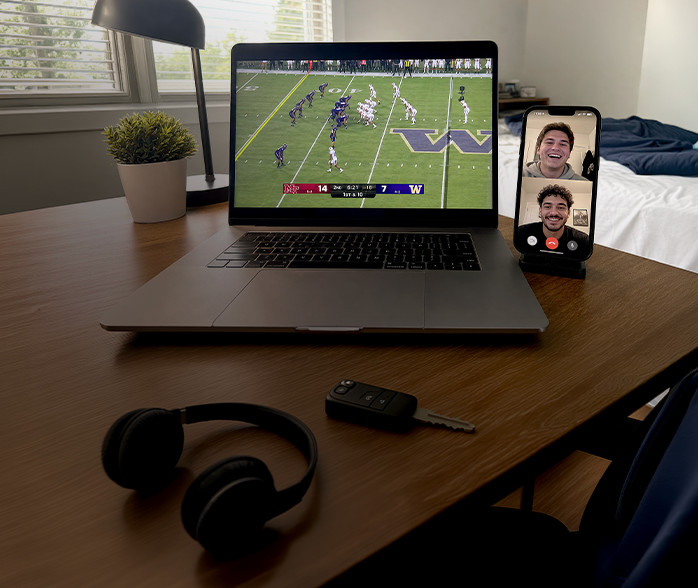 Computadora portátil que muestra un partido de fútbol americano junto a un teléfono inteligente en videollamada con dos hombres sonrientes, sobre un escritorio