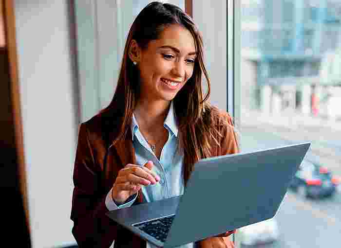 A professional woman in a blazer stands by a window overlooking a city while working on her laptop using Optimum Business Internet.
