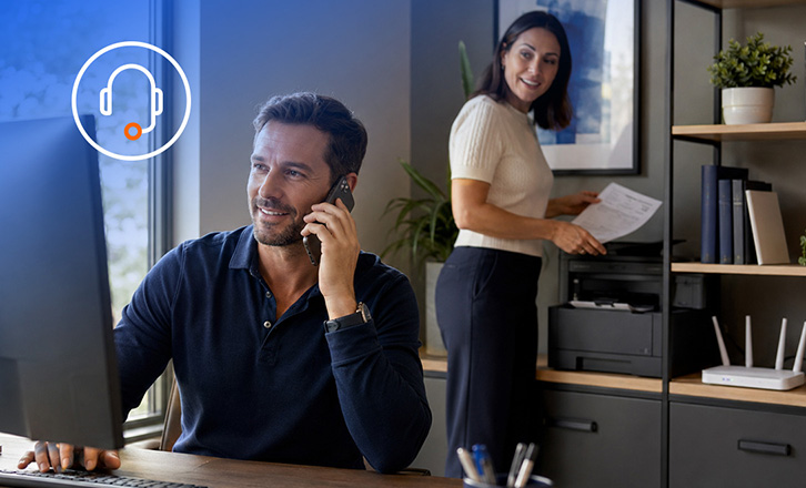 Man on a phone call sitting in an office at a desktop computer in front of a woman standing holding documents 