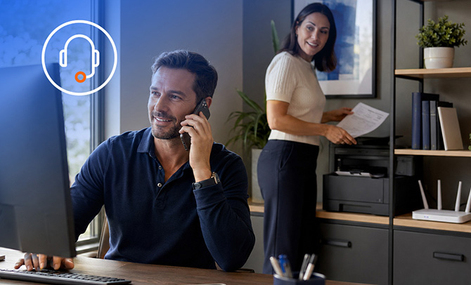 Man on a phone call sitting in an office at a desktop computer in front of a woman standing holding documents 