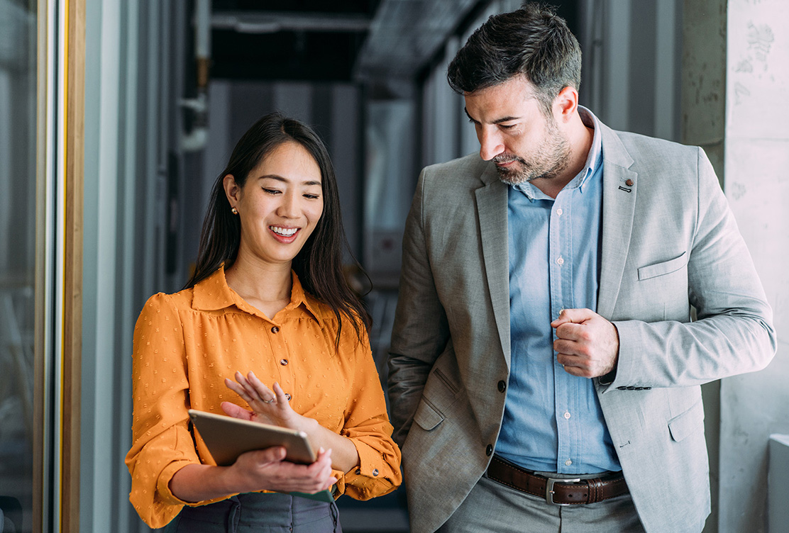 Two colleagues in business casual attire walking and talking together in a modern office hallway 