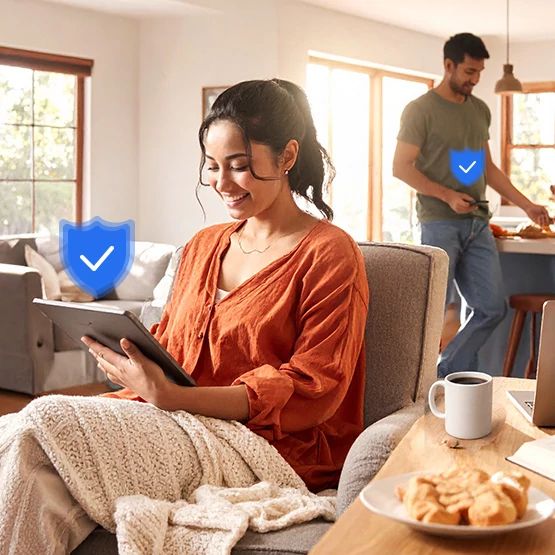 Mujer con cárdigan color óxido sentada en un sofá usando un teléfono inteligente, hombre relajándose detrás de ella, comida sobre la mesa, insignia de verificación azul