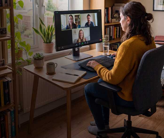 Woman in mustard sweater on a video call with three colleagues on a desktop monitor at a wooden desk