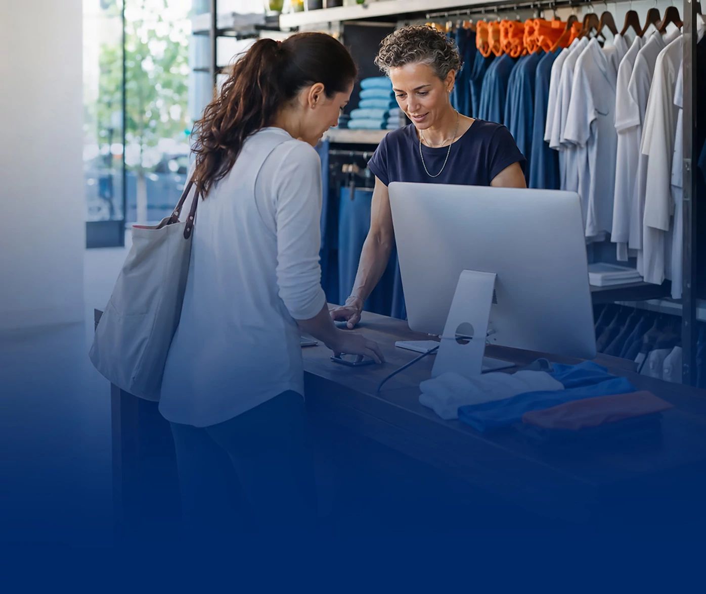 Two women looking at a desktop computer screen inside a clothing boutique with garment racks in the background