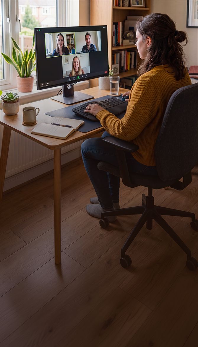 Woman in mustard sweater on a video call with three colleagues on a desktop monitor at a wooden desk