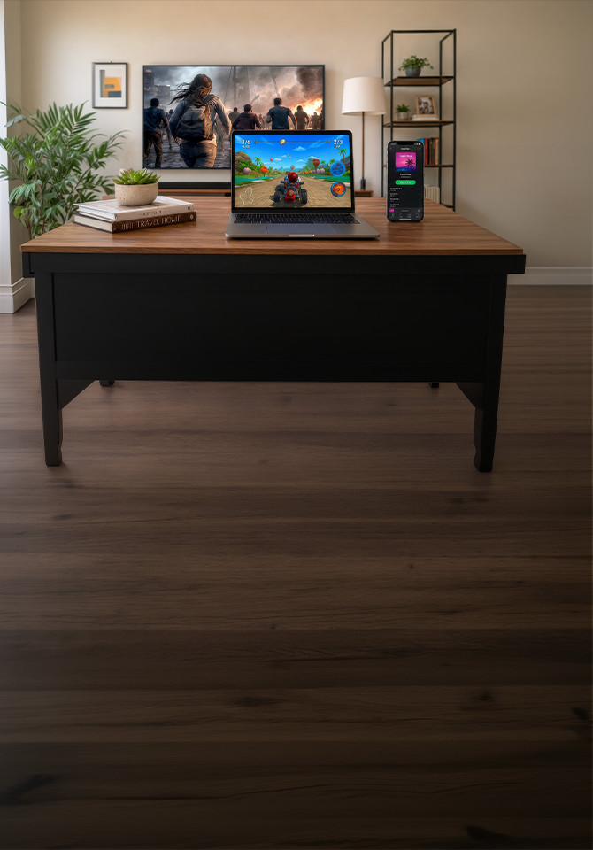 Mesa de centro en la sala de estar con un teléfono inteligente y una computadora portátil que muestran un juego de carreras, frente a un televisor que reproduce una escena de acción