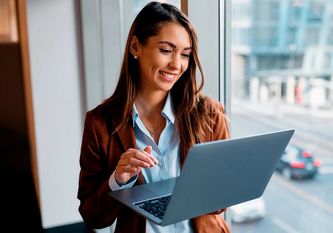 A professional woman in a blazer stands by a window overlooking a city while working on her laptop using Optimum Business Internet.