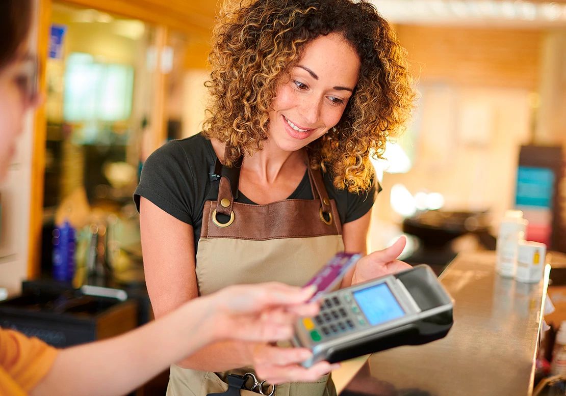 A small business owner smiles while processing a customer's credit card payment on a handheld terminal using Optimum Business Internet.