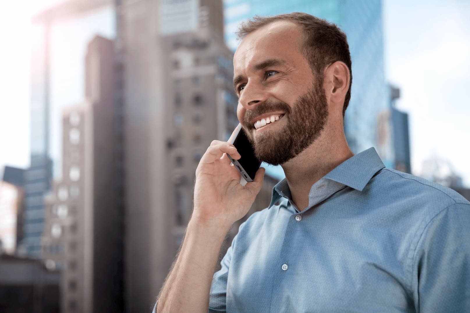 A young man with glasses and a beard smiles while using a new smart phone as he leans against the sales counter of a technology retailer