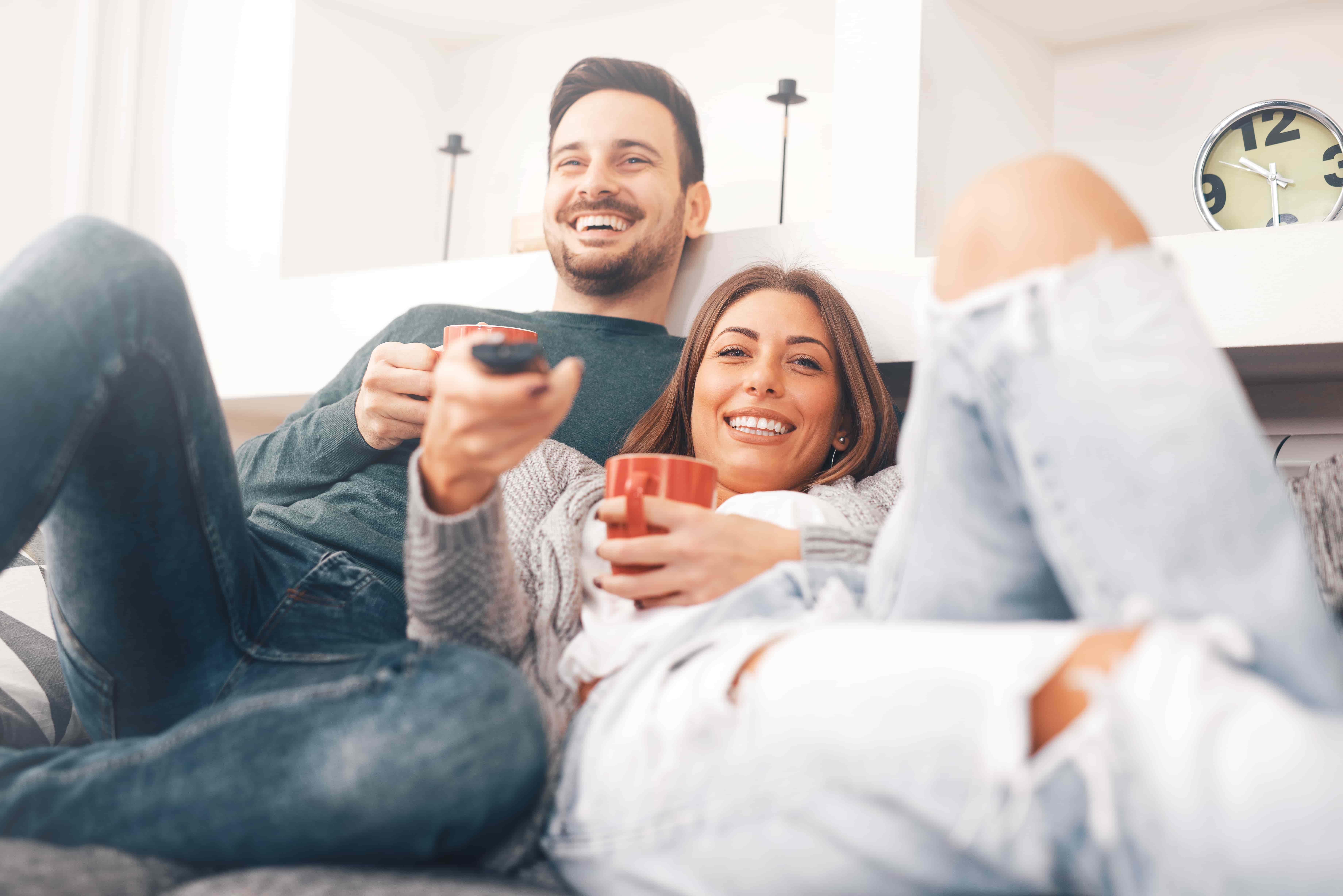 A young couple reclines while smiling on their couch, with remote control poised to choose which TV program to watch