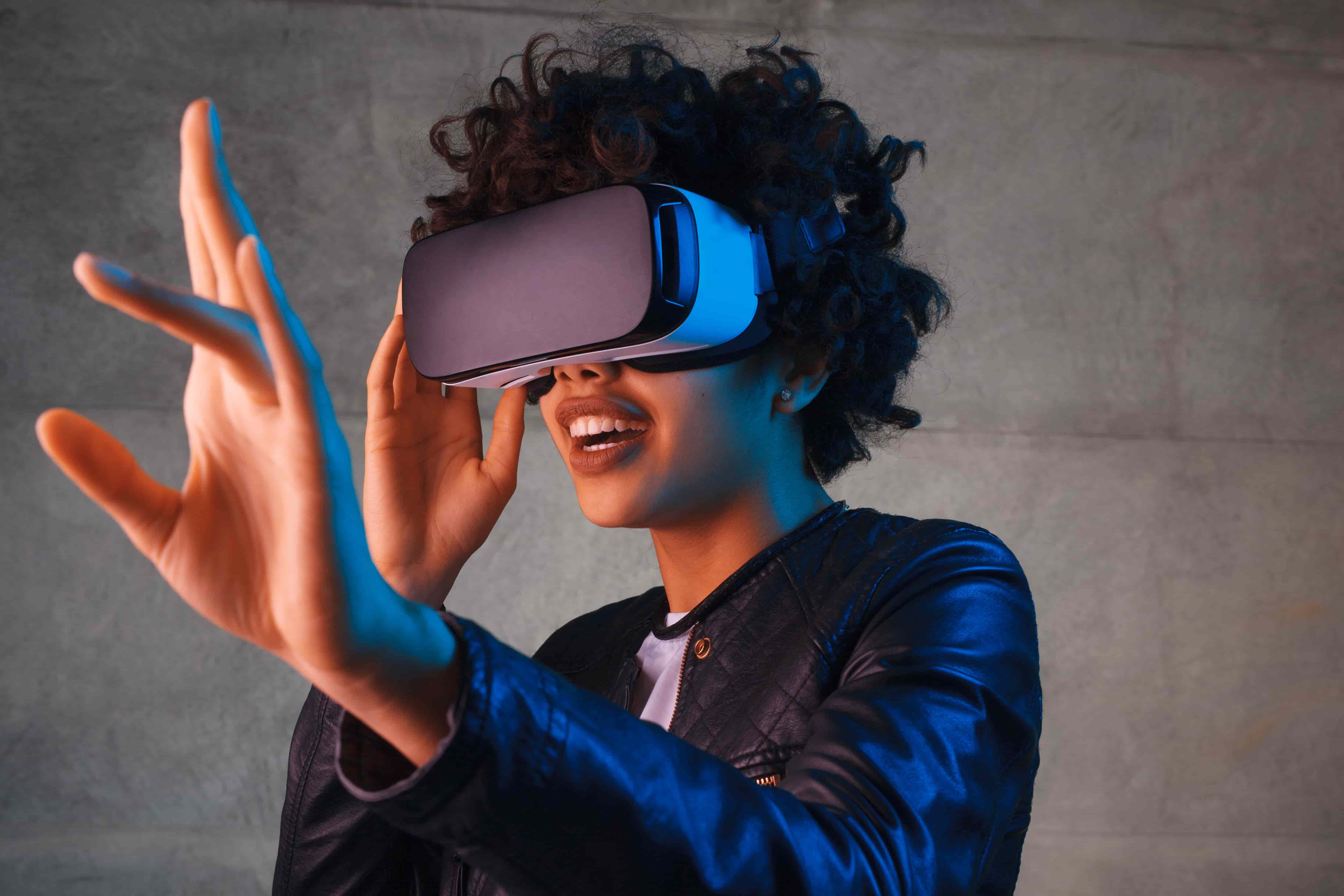 A young African American woman wearing a virtual reality headset holds her hand out in front of her as if touching an unseen object.