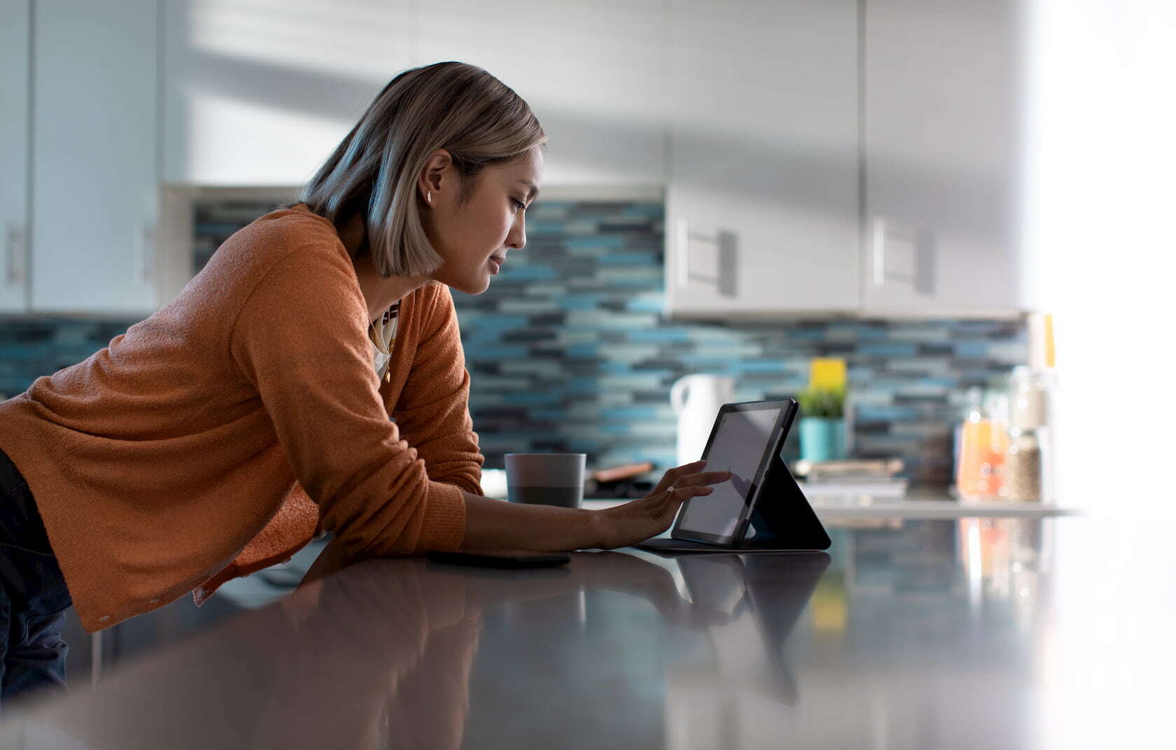 A young woman in her brightly lit kitchen leaning on her counter in the early morning, enjoying her coffee and tablet connected over wifi