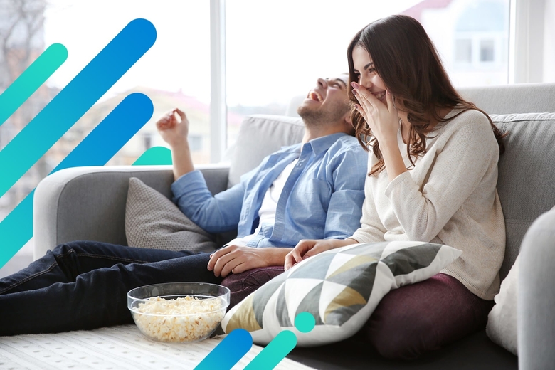 A couple laughs hysterically while relaxing on a sectional couch, watching TV with a bowl of popcorn sitting between them.