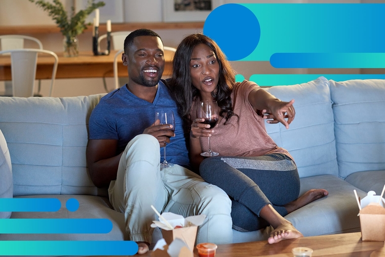 An African American couple smiles while watching a movie on their couch, each is holding a glass of red wine